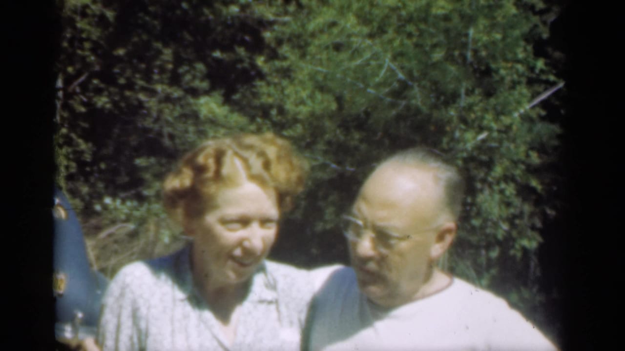 Older woman smiles while an older man with glasses looks toward her outdoors.