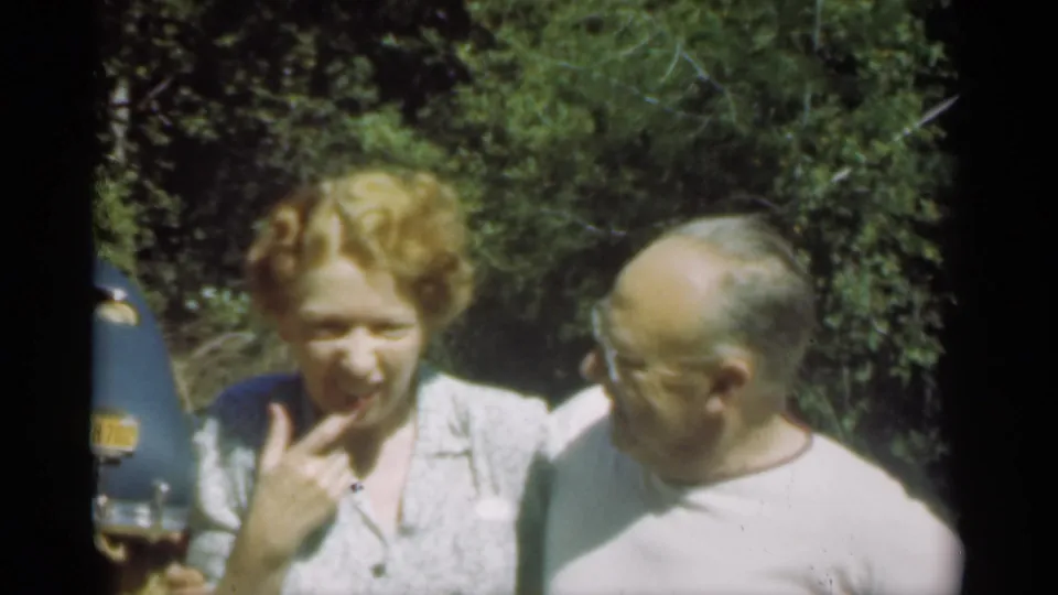 The older woman gestures near her mouth while the older man watches her, with trees and part of a blue car behind them.