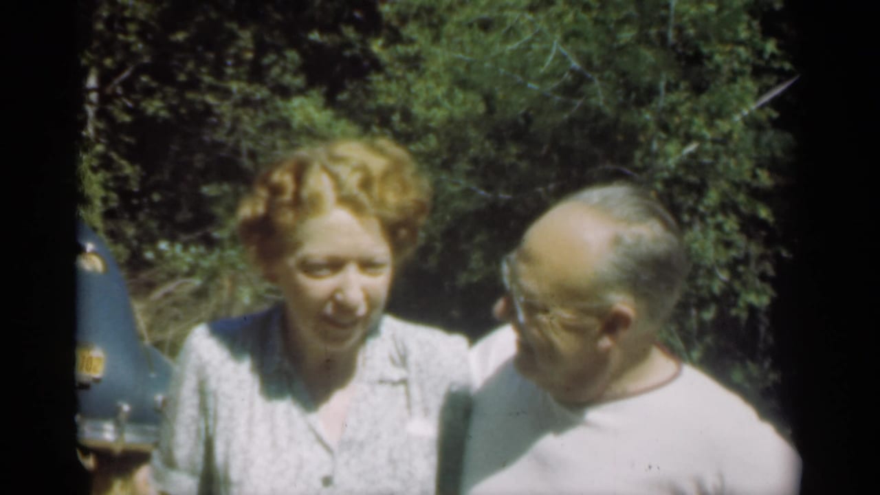 Two adults outdoors, a red-haired woman facing camera and a man in glasses turned toward her.