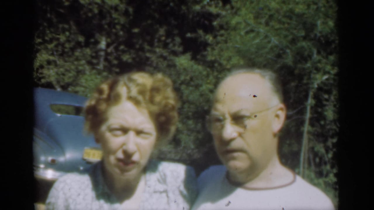 Adult couple standing outdoors near a blue car with trees behind them.