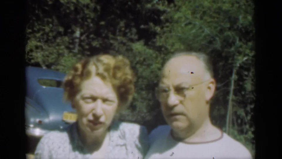 In Santa Cruz home-movie footage from 1949, an adult couple stands in bright sun beside a blue car and wooded background.