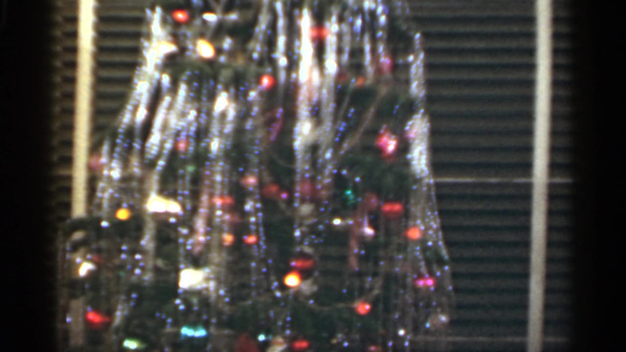 Decorated Christmas tree with tinsel and colored lights seen through horizontal blinds.