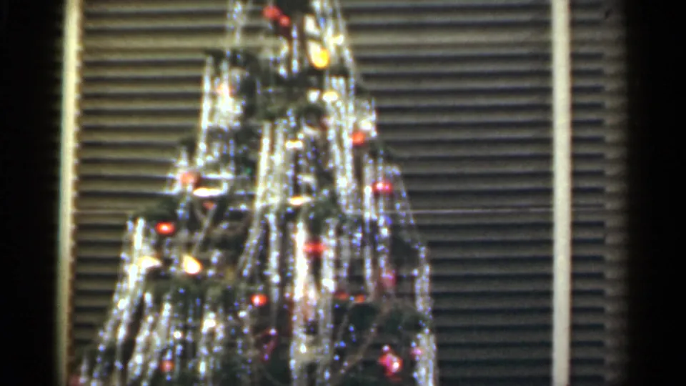 Santa Cruz 1949 8mm frame of a decorated Christmas tree, with bright tinsel strands, colored ornaments, and blinds visible behind the tree.