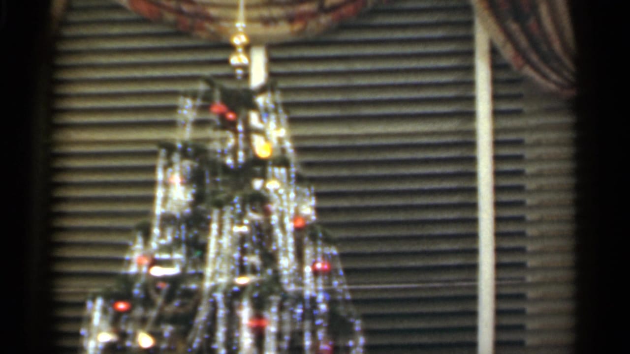 Tinsel-covered Christmas tree with glowing lights in front of venetian blinds.