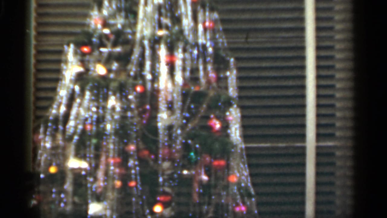 Tinsel-covered Christmas tree in front of window blinds.