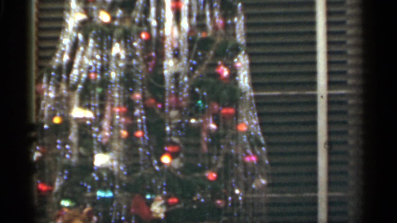 Decorated Christmas tree with tinsel and colored lights before blinds.