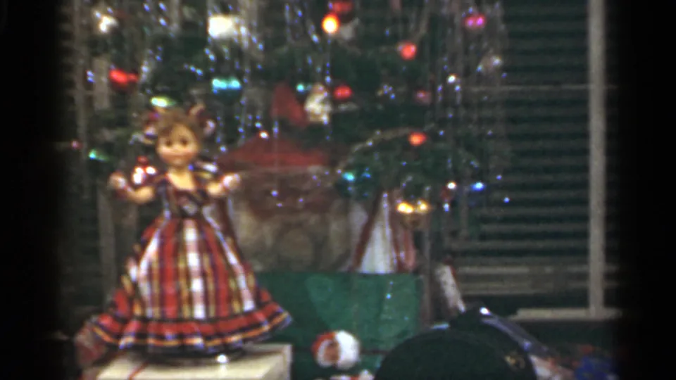 1949 Santa Cruz home-movie frame of a plaid-dress doll standing beneath a tinsel Christmas tree among wrapped gifts.