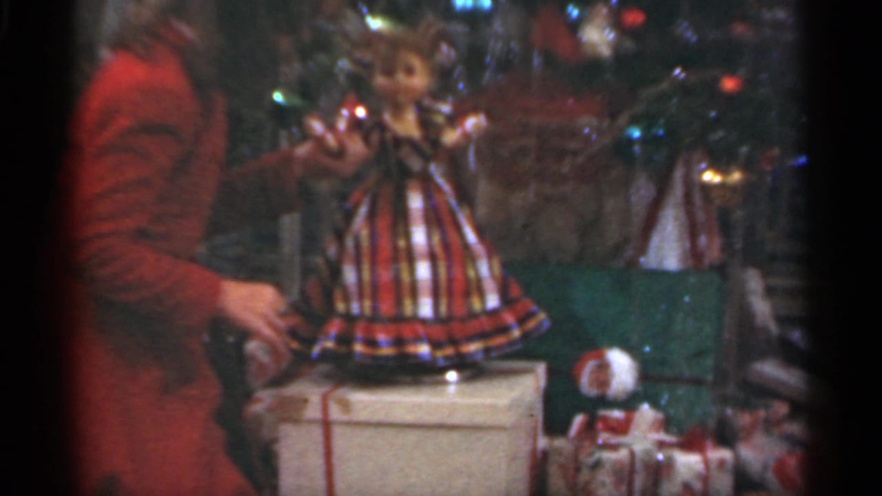 Girl in a red dress holds a doll beside a Christmas tree and wrapped presents.