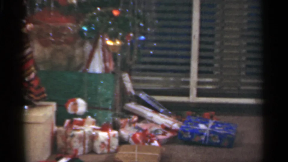 A 1949 home-movie frame shows Christmas packages, a small doll, tree tinsel, and venetian blinds in a private room.