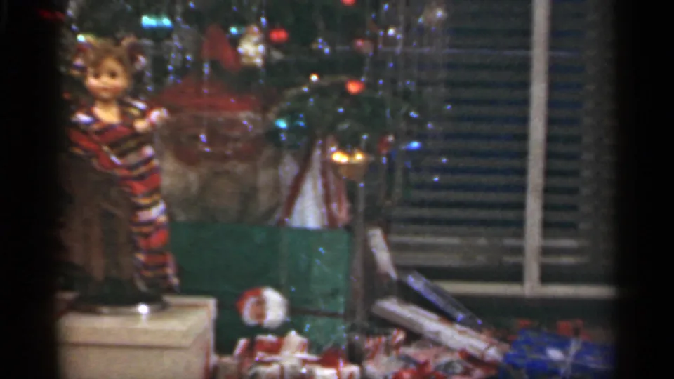 The frame centers on a plaid-dressed doll beside the Christmas tree, with gifts and blinds visible in the Santa Cruz interior.