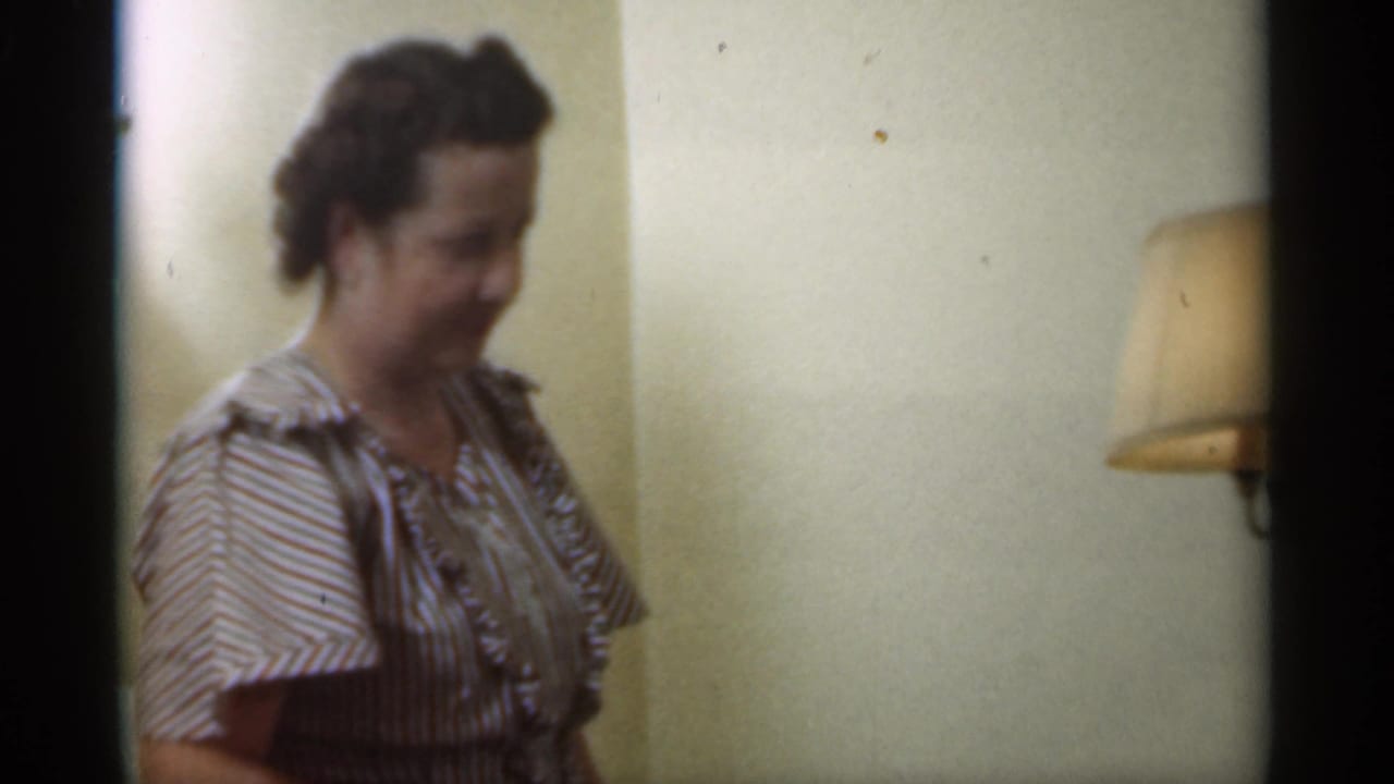 Adult woman in a striped dress standing indoors near a shaded wall lamp.