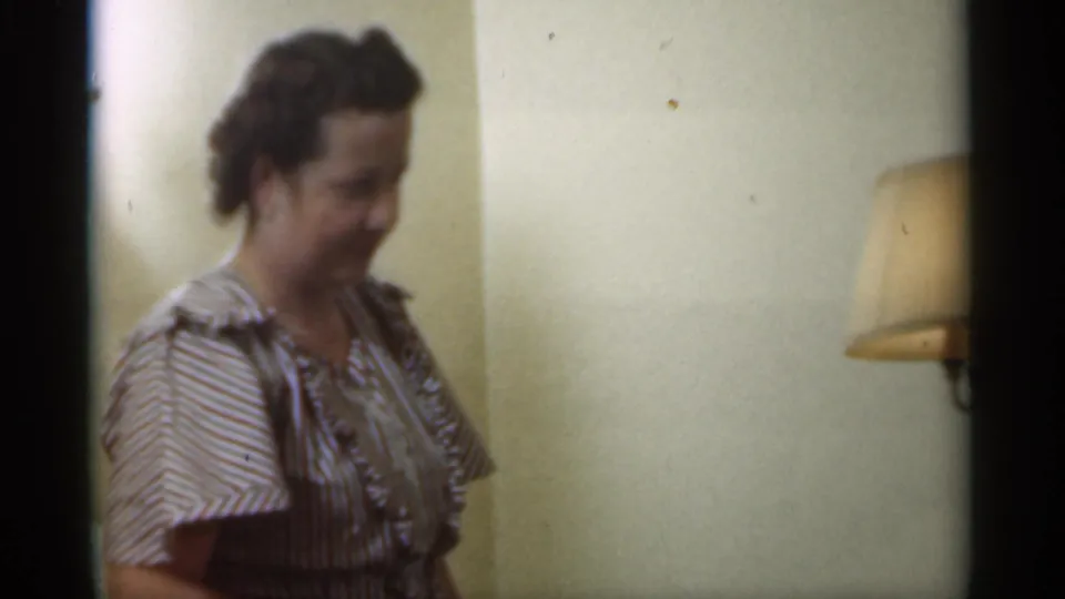 Adult woman in a striped dress stands in a Santa Cruz home interior in 1949, with a shaded wall lamp beside her.