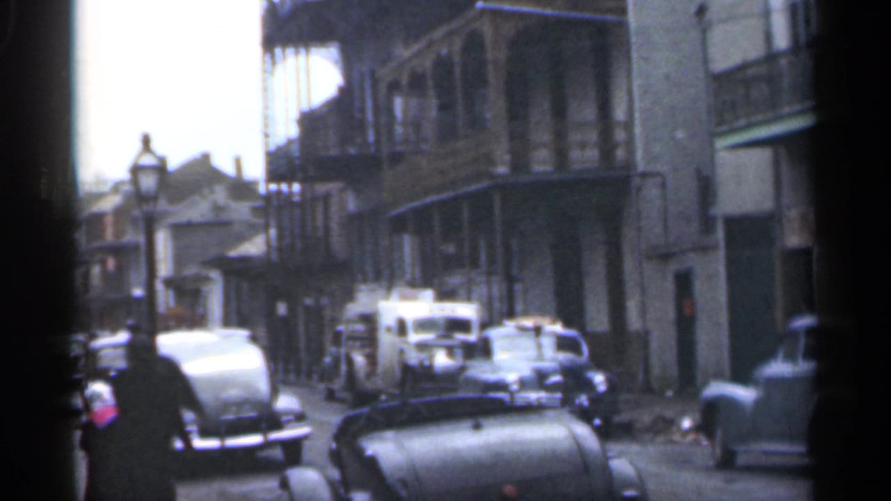 Parked cars line a narrow street as a woman walks past buildings with balconies.