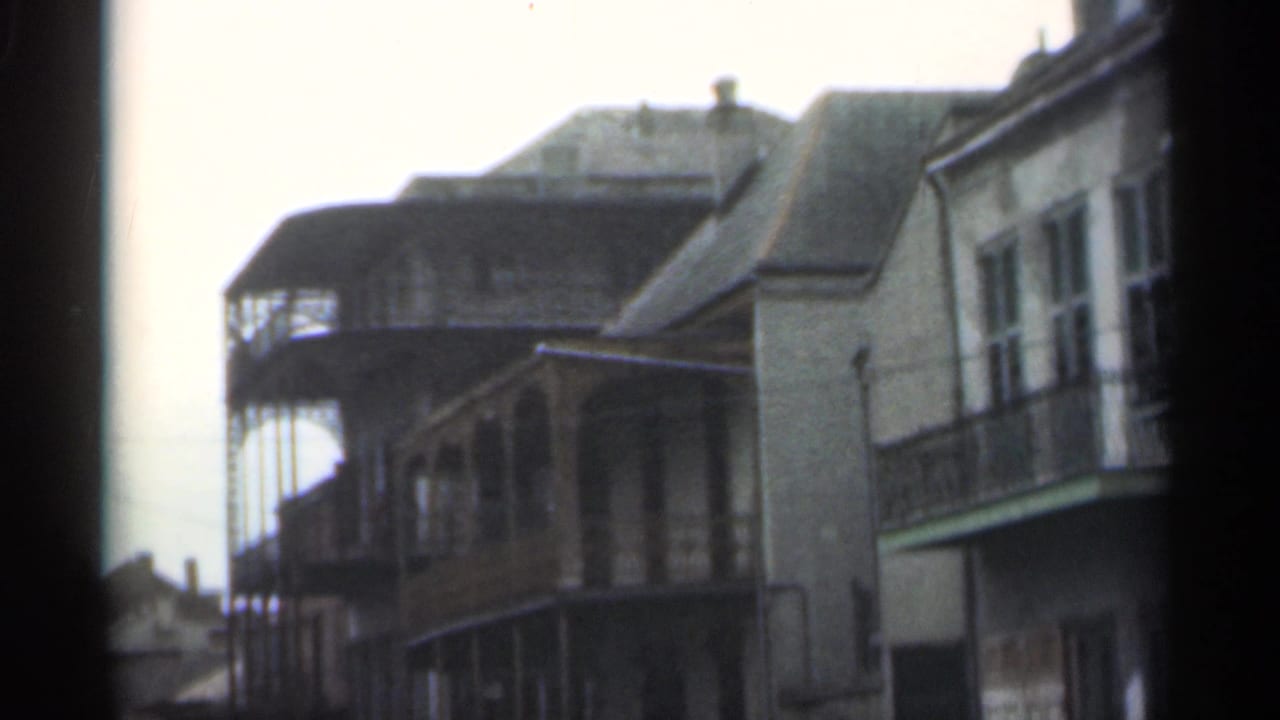 Two-story street buildings with balconies and a pale sky, seen from a soft handheld 8mm frame.