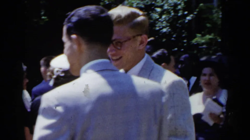 1954 New York wedding guests mingle outdoors, with men in pale suits, ties, and boutonnieres visible in leafy shade.