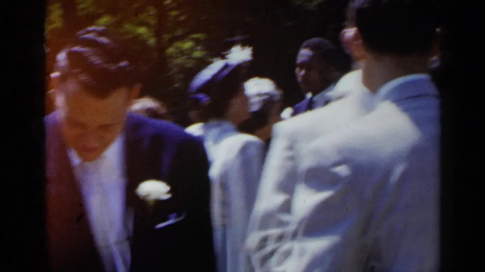 At an outdoor 1954 New York wedding, a man in a dark suit and white boutonniere stands amid guests in formal attire.