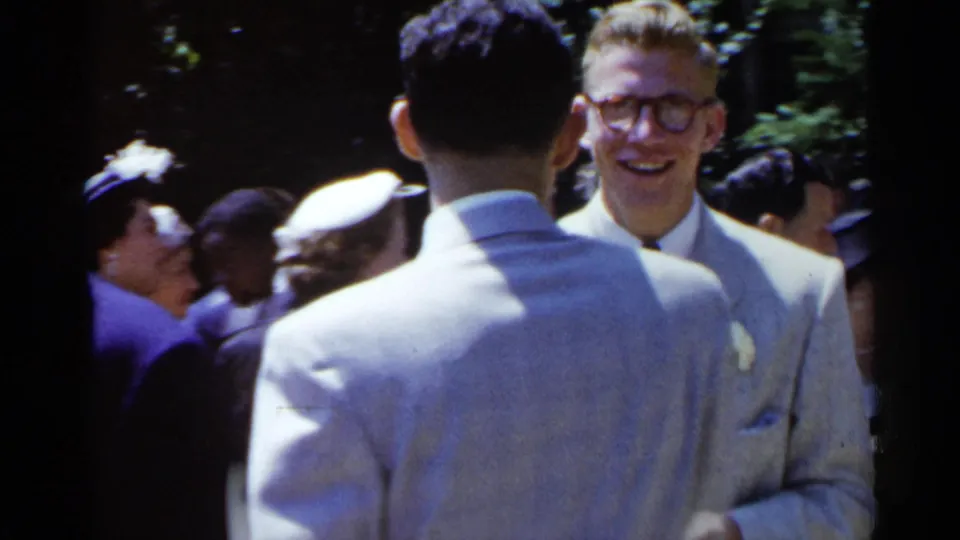 Two pale-suited men stand face to face at a 1954 New York outdoor wedding, with smiling guests and hats visible behind them.