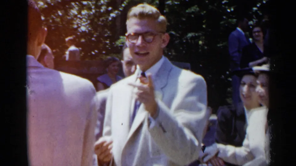 A man in a light suit and glasses gestures at a 1954 New York wedding, with a seated woman in white partly visible at the edge.