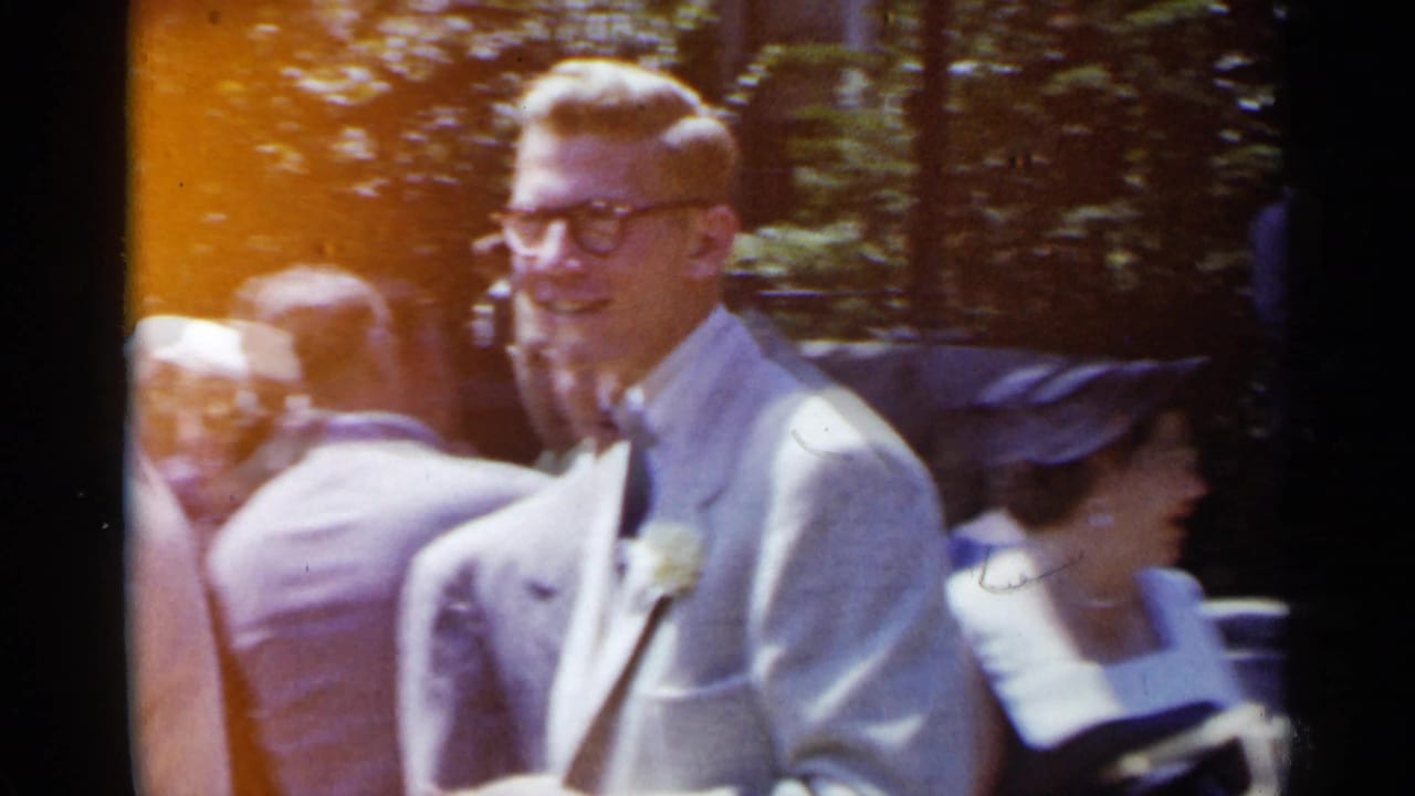 Man in a light suit and glasses stands among formally dressed guests outdoors.