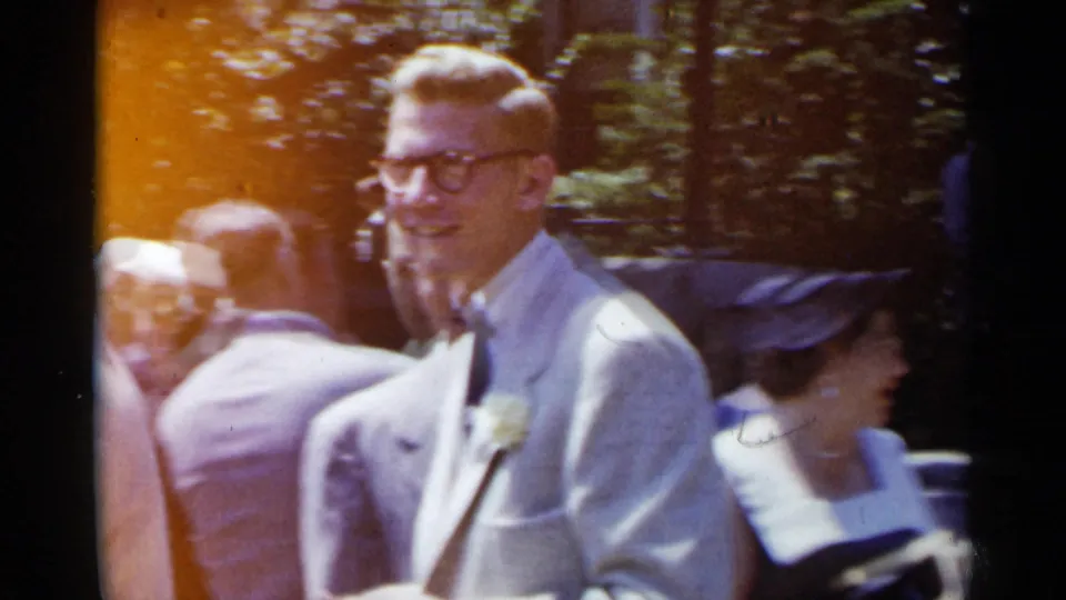 1954 New York home-movie frame showing a man in a light suit and glasses among guests at a sunlit formal outdoor gathering.