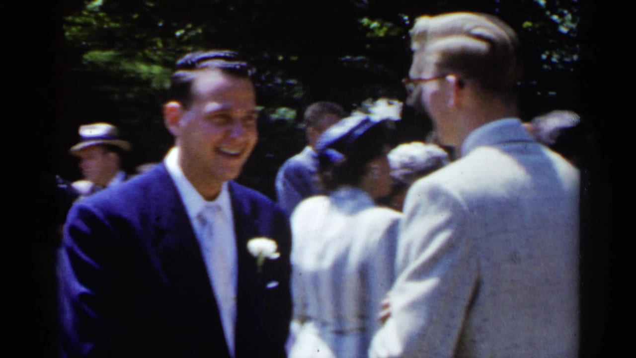 Smiling man in a dark suit faces another man in a light suit outdoors.