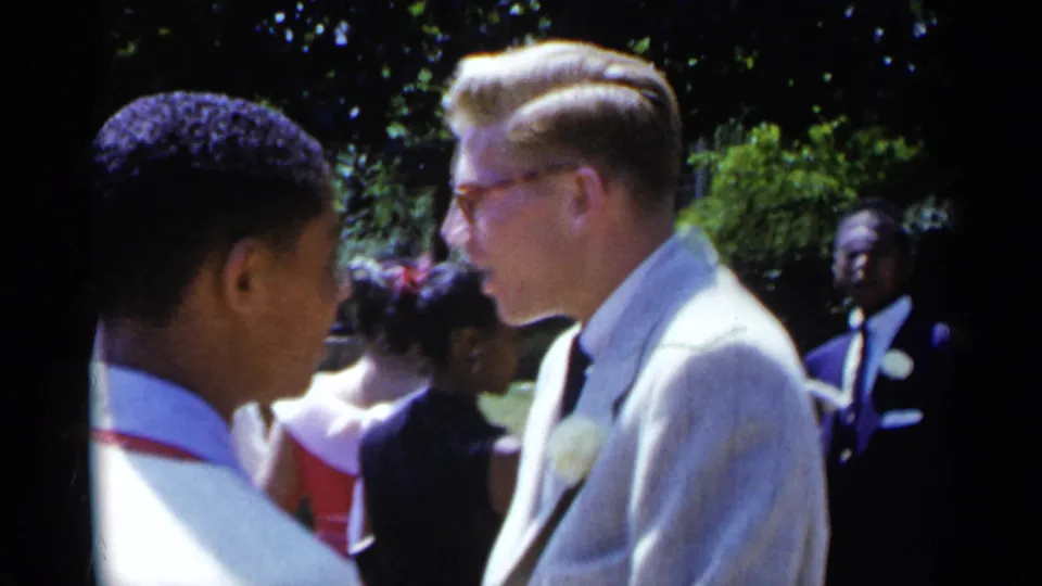 Two young Black men in white shirts and red suspenders stand close to a man in a light suit at the outdoor gathering.