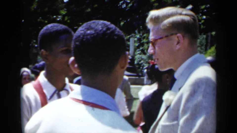The camera frames two young Black men from behind as they face a man in a light suit at the 1954 New York gathering.