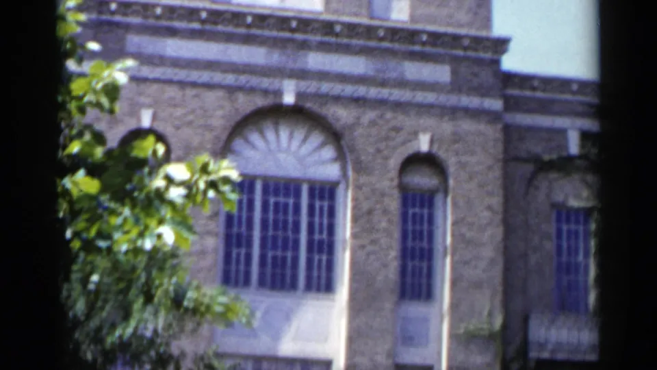 1954 New York 8mm frame of an ornate building facade with a large arched window, stone detailing and foreground foliage.