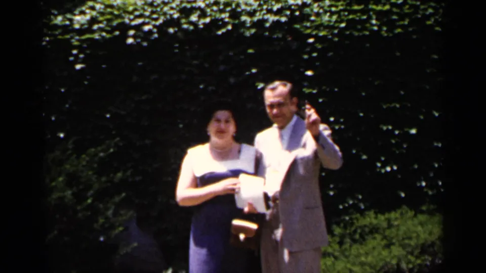 In the supplied 1954 New York home-movie setting, an adult couple stands in a garden, the man in a light suit and the woman in a dark dress with a white top.