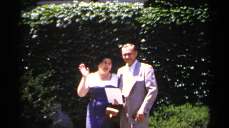 The woman waves toward the camera while standing with a suited man in the garden, consistent with the supplied 1954 New York home-movie metadata.