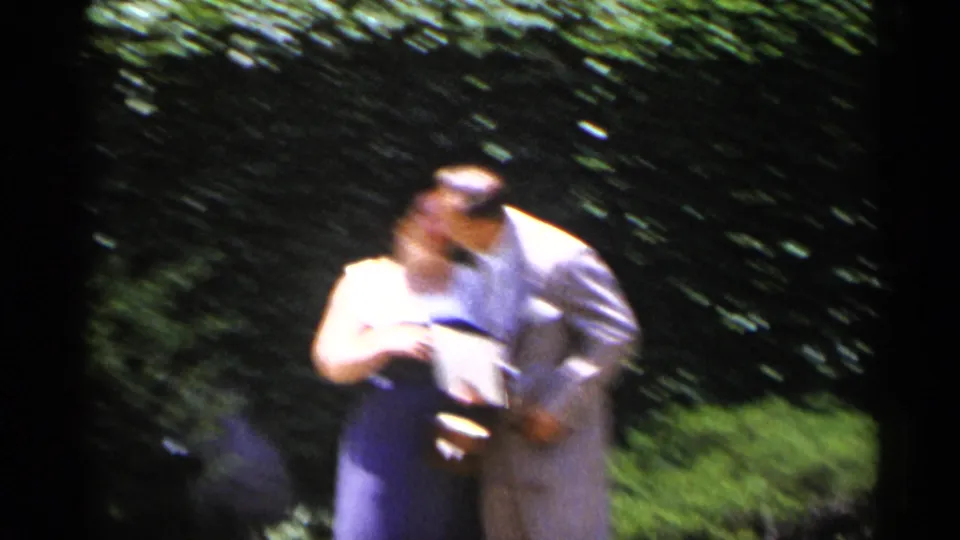An adult couple kisses in the garden, the man in a light suit and the woman in a dark dress with a white top, recorded with soft handheld 8mm motion blur.