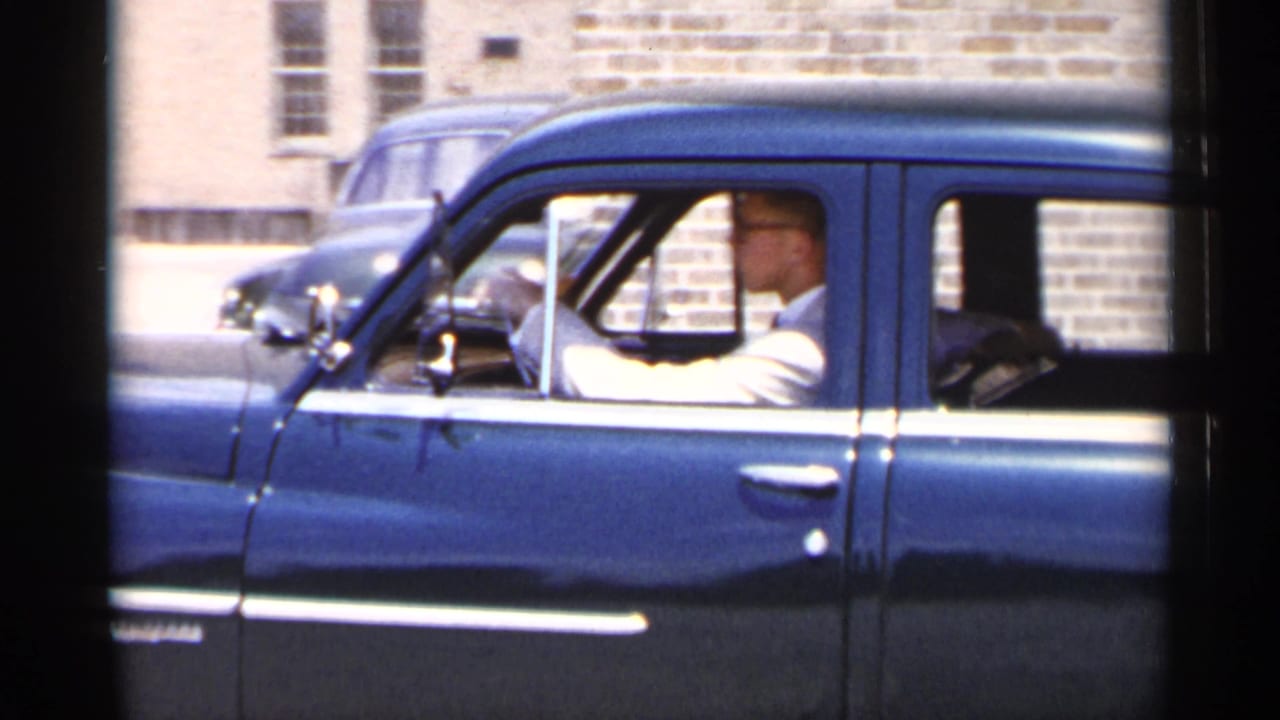 1954 New York home-movie frame showing a blue mid-century sedan in profile with a man in glasses driving past brick buildings.
