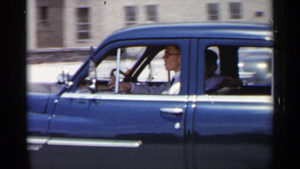 1954 New York frame of a blue sedan moving past the camera, with the driver at the wheel and a rear passenger in shadow.