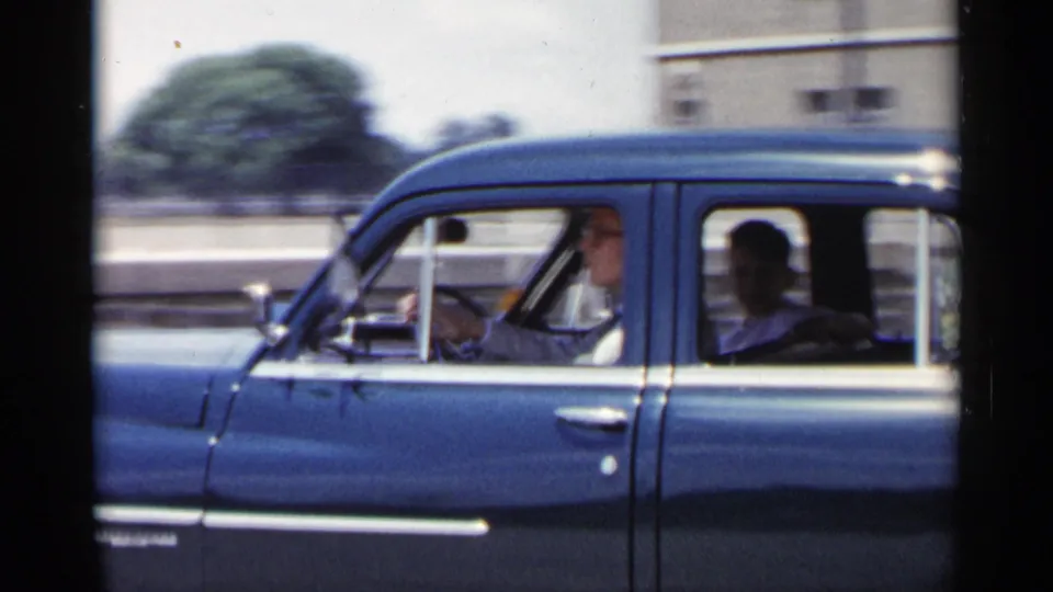 1954 New York home-movie frame showing the blue sedan closer to the camera, with the driver and a rear-seat passenger visible as the background blurs.