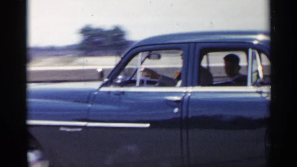 Late-frame view of the 1954 New York blue sedan continuing along the street, with occupants seen through the side windows and a soft urban roadside behind it.