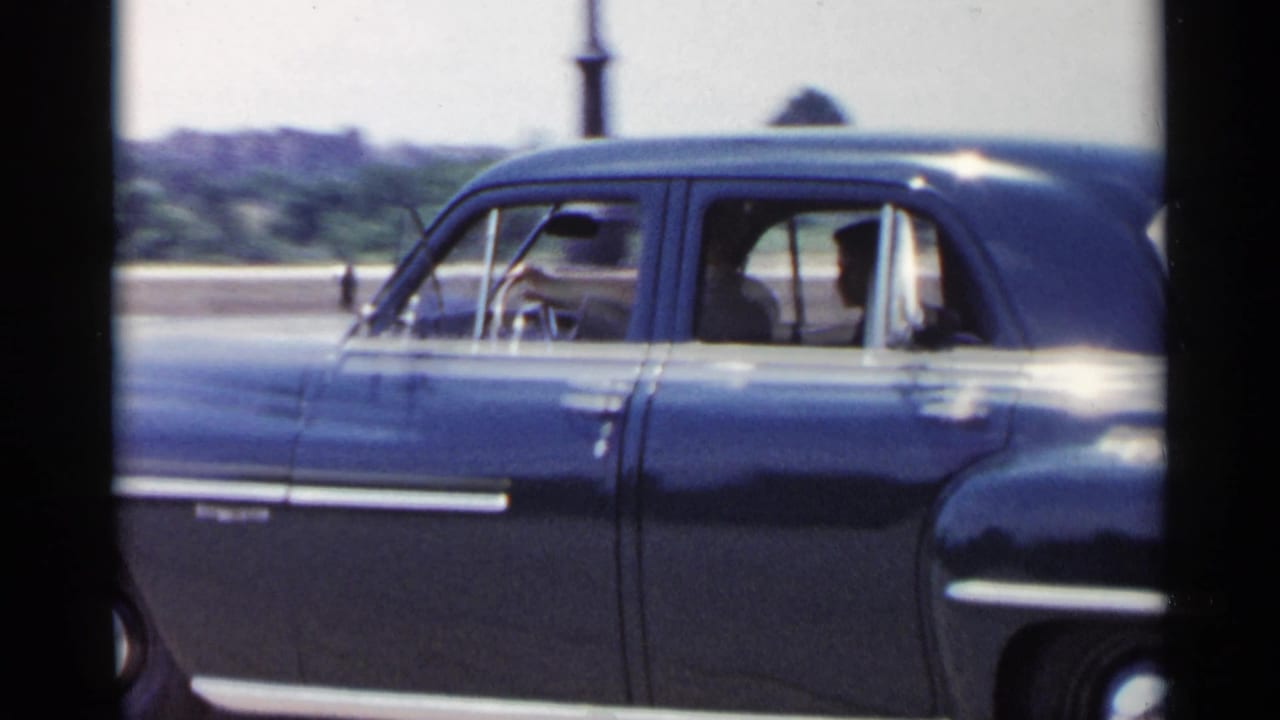 Black mid-century sedan seen from the side with occupants visible through open windows.