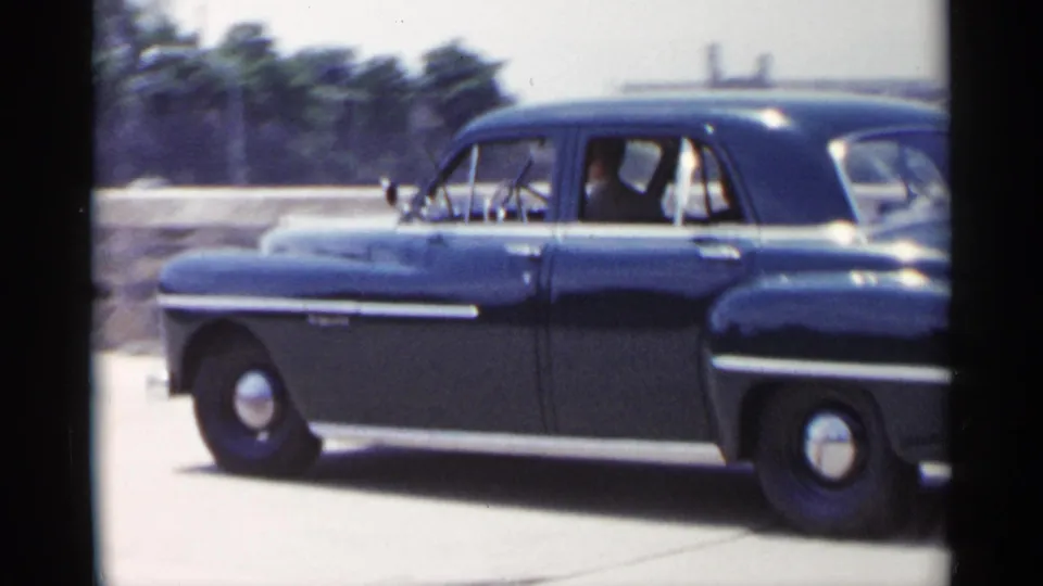 Side view of a black mid-century sedan moving through a New York street, with occupants visible in the front and rear seats.