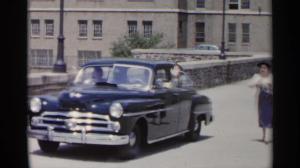 Front three-quarter view of a black 1950s sedan near a stone wall and large building, with a woman in period dress on the sidewalk.
