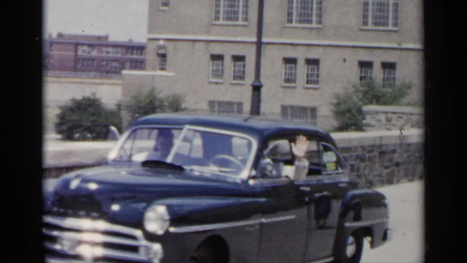 1954 New York frame showing a passenger waving from a black sedan beside a stone wall, sidewalk, street lamp, and large building.