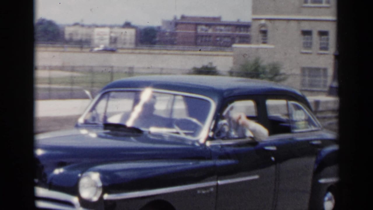 Dark blue mid-century sedan passing on a city street with a driver visible through the front window.
