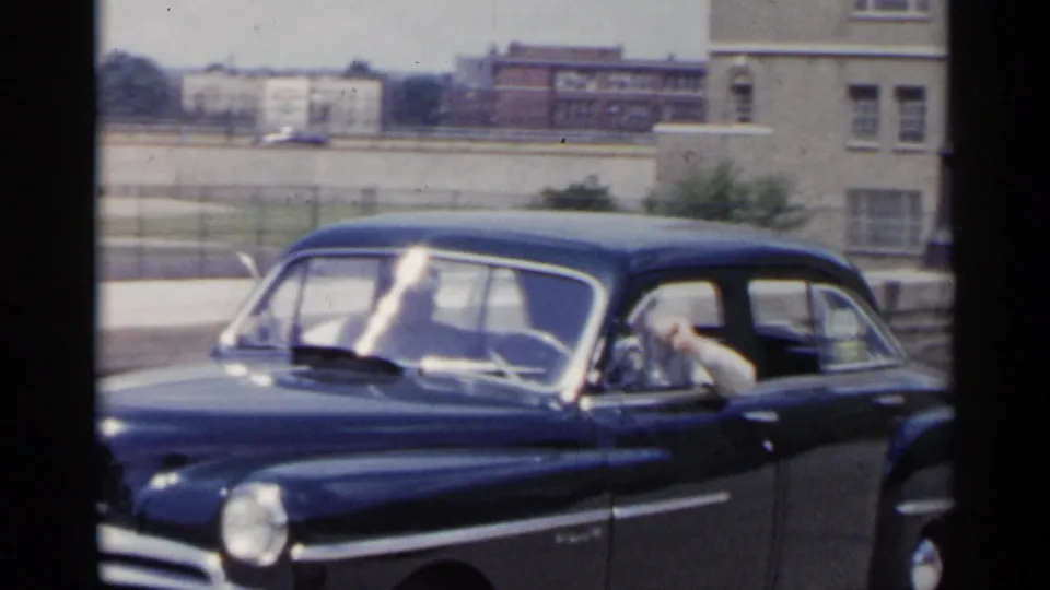 1954 New York home-movie frame showing a dark blue sedan on a city street, with a driver visible and buildings in the background.