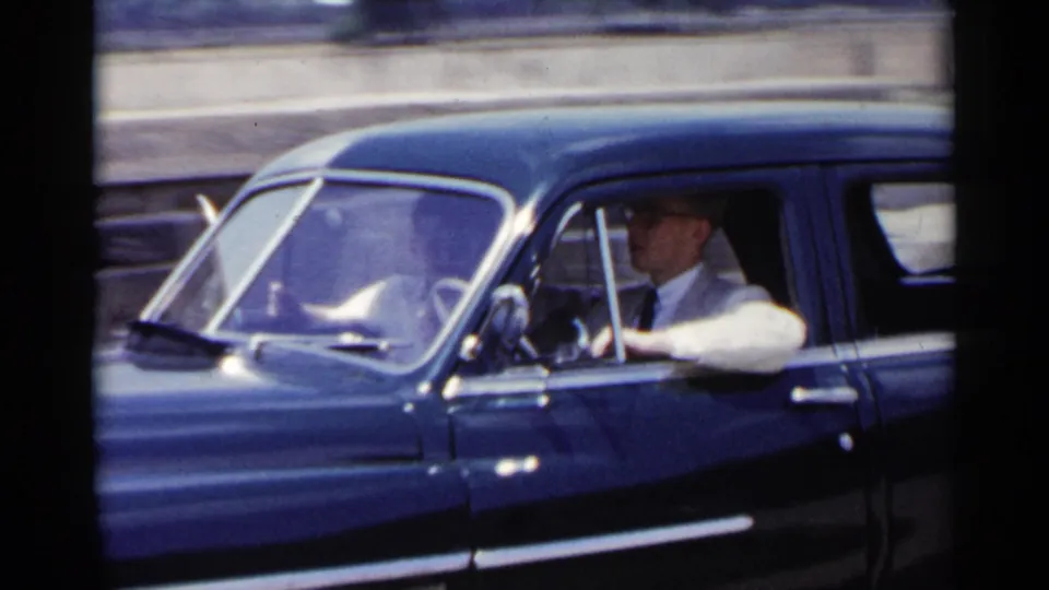 Close 1954 New York frame of a man in a light jacket and glasses driving a dark blue mid-century sedan.