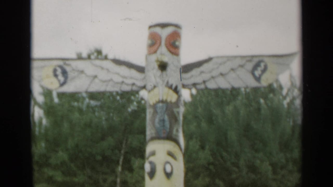 Painted totem-style pole with wing-like carvings in front of trees.