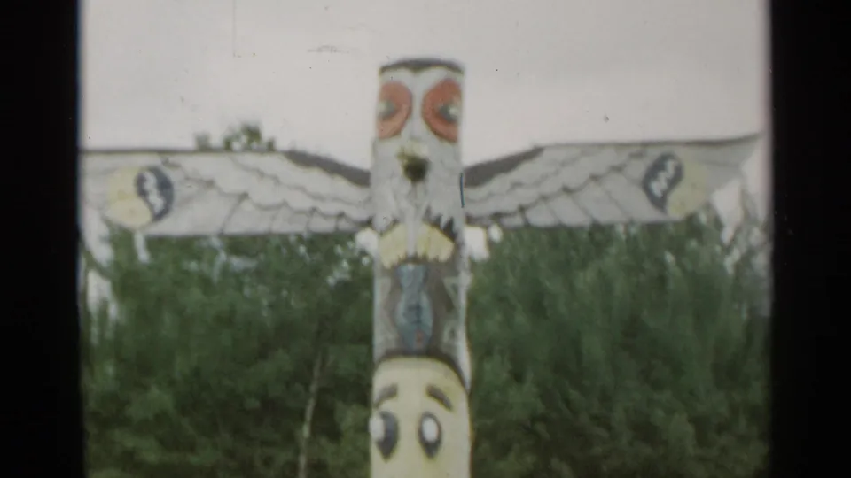 1954 New York 8mm frame showing the upper section of a painted totem-style pole with wing-like carvings against dense trees.