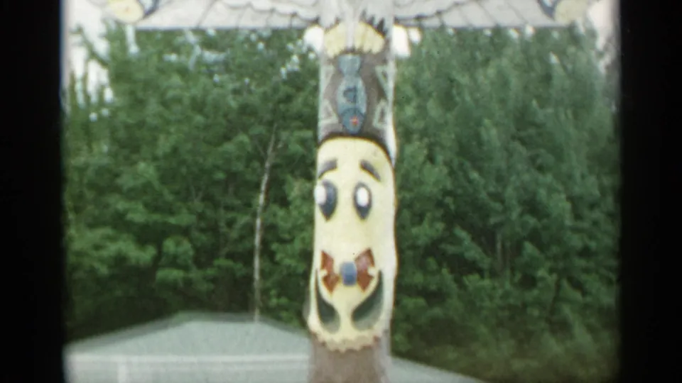 1954 New York 8mm frame with a painted totem-style pole, stylized face details, trees, and a small light-colored roof in the background.