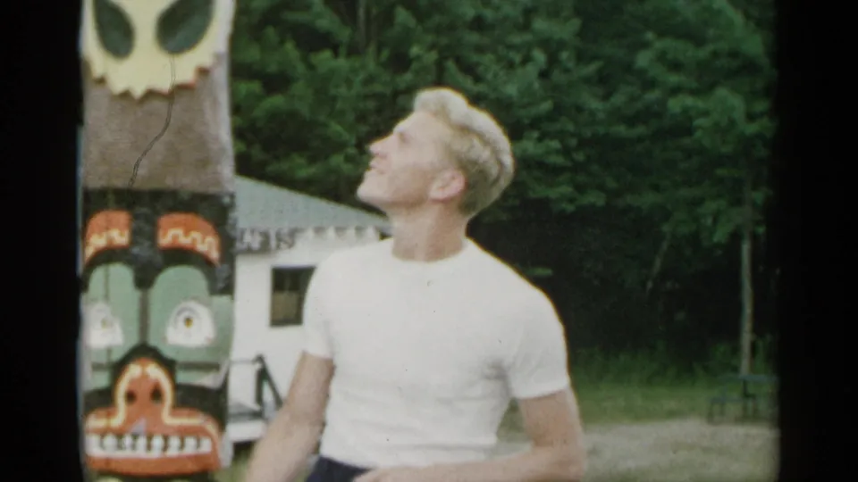 1954 New York 8mm frame showing a young man in a white T-shirt beside a painted totem-style pole, looking upward in a garden setting.