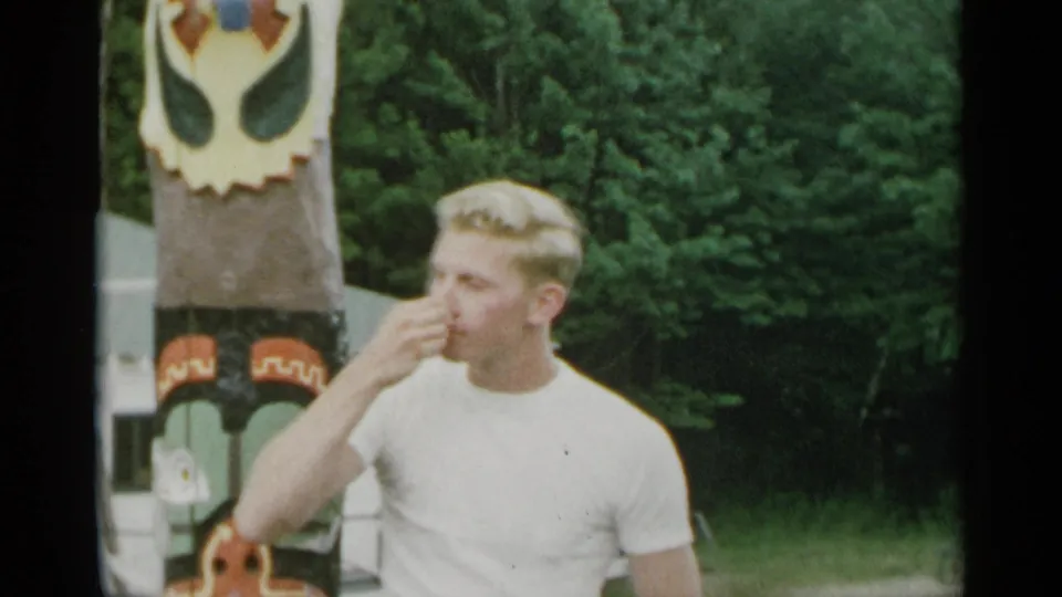 1954 New York 8mm frame of a young man beside a painted totem-style pole, raising his hand to his mouth with trees behind him.