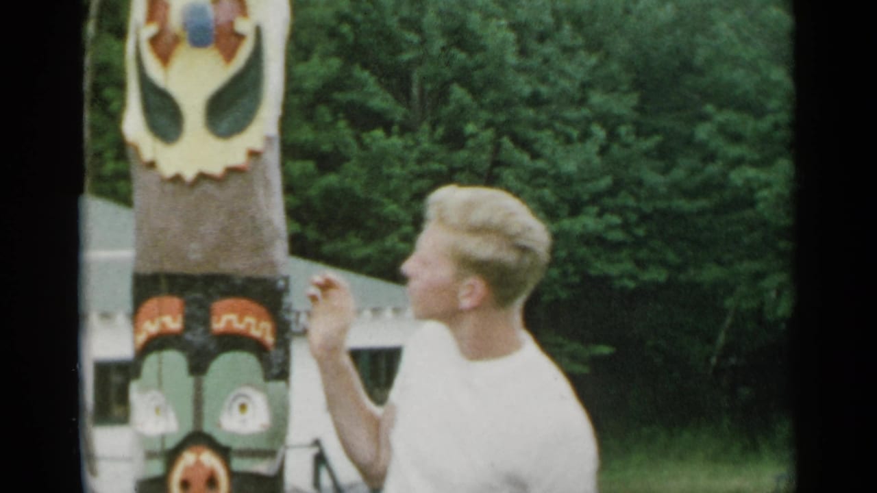 Man in a white T-shirt stands beside a painted totem pole outdoors.