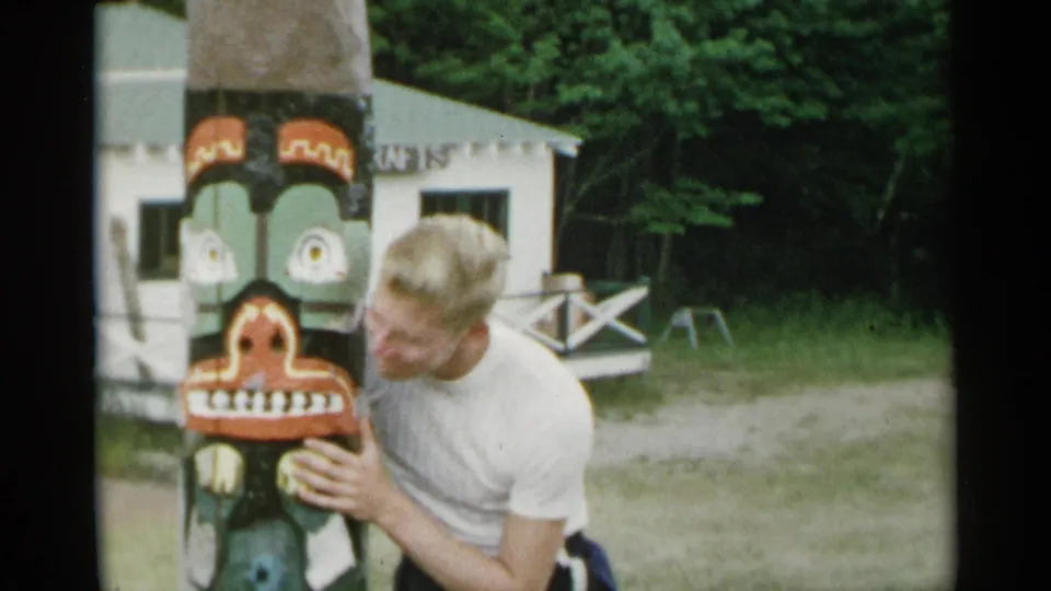 The man leans in close to the painted totem pole, holding it with both hands in the outdoor New York scene.
