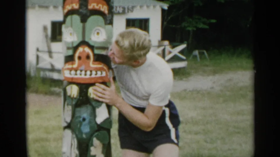The man bends toward the painted face on the totem pole, with a white building and wooded background behind him.